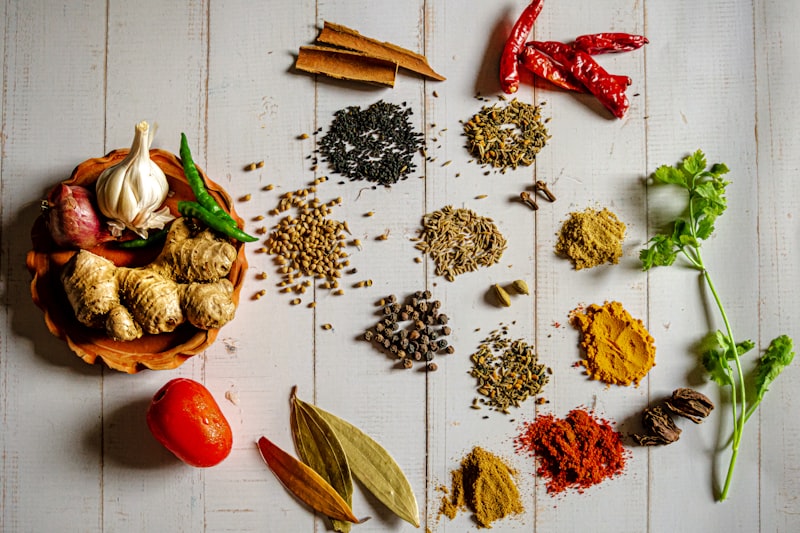 A flat-lay arrangement of whole spices, dried herbs, ginger, garlic and chilli peppers on a white wooden surface