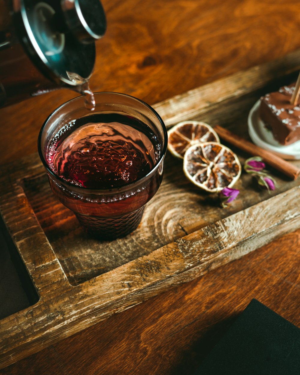A hot glass of red-amber Persian chai being poured from a metal teapot on a wooden tray with dried citrus and rose